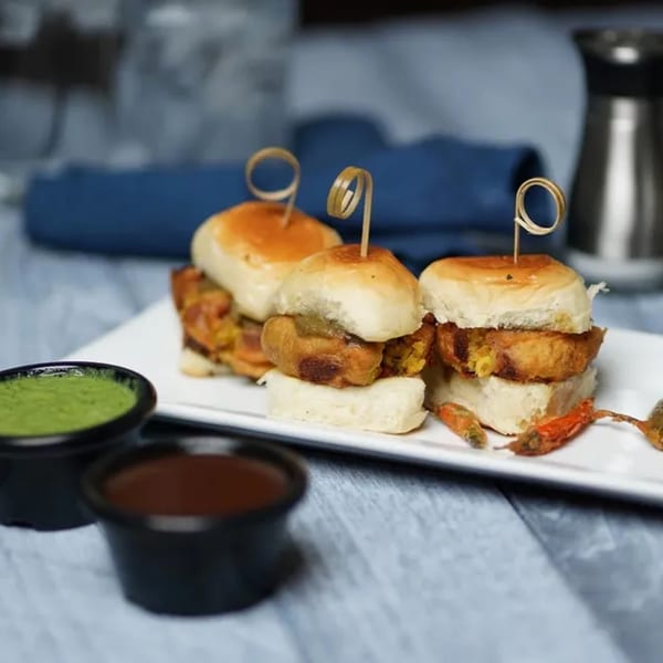 Three Indian vada pav sliders with green and brown chutneys on a white plate.