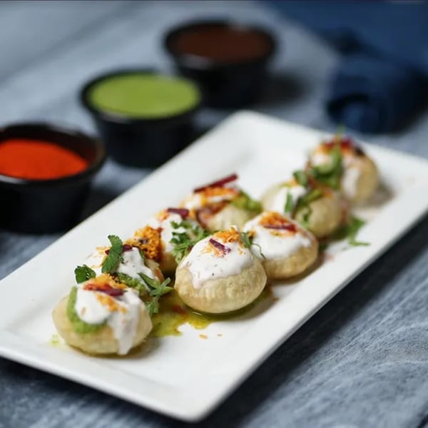 A plate of Clay Oven's Indian Dahi Puri, topped with yogurt, spices, and herbs, with sauces in the background.