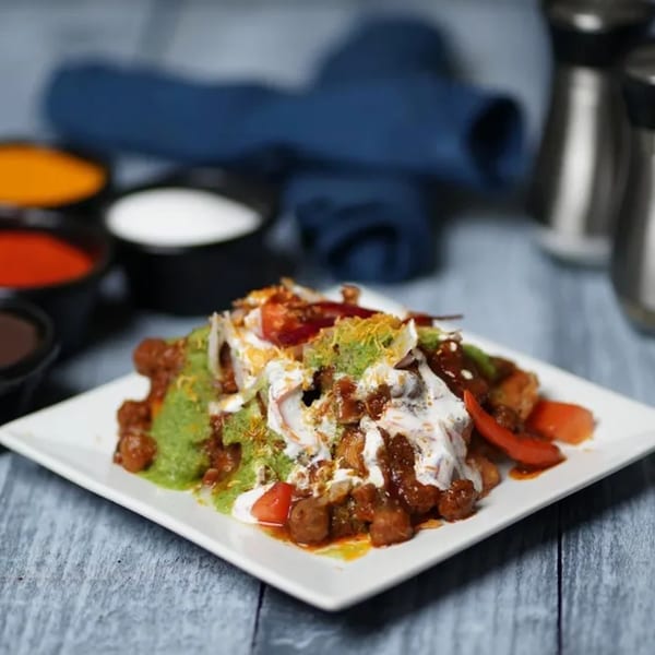 A vibrant Indian chaat dish, topped with green chutney, yogurt, and fresh vegetables, on a white plate.